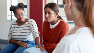 Image of 3 women in a group therapy type environment. The woman in th center is crying while the other two offer comfort.
