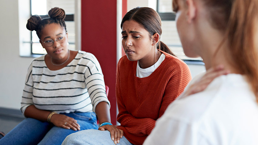 Image of 3 women in a group therapy type environment. The woman in th center is crying while the other two offer comfort.