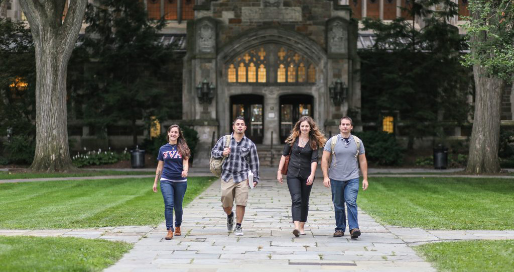 4 Student Veterans walking across a paved sidewalk in front of a campus building at a University.